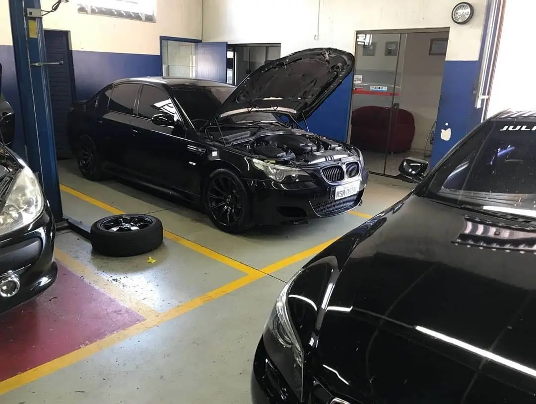 BMW preta com o capô aberto na oficina, ao lado de outros carros importados